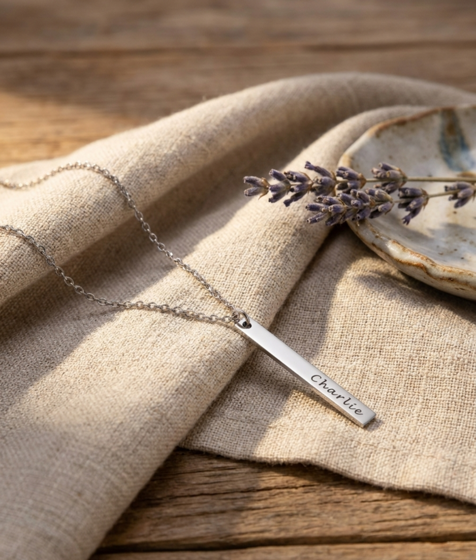 Zilveren rechthoekige staafvormige hanger met naam gravure aan zilveren ketting liggend op linnen doek met lavendel lifestyle modelfoto gepersonaliseerd zilveren naamketting sieraad